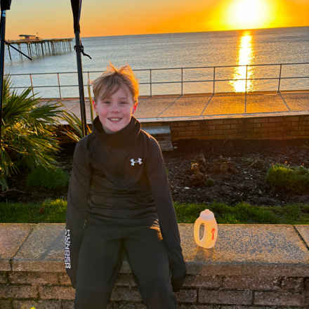 Picture shows young 10 year old Archie sitting on a wall at the coast with sunset in the background