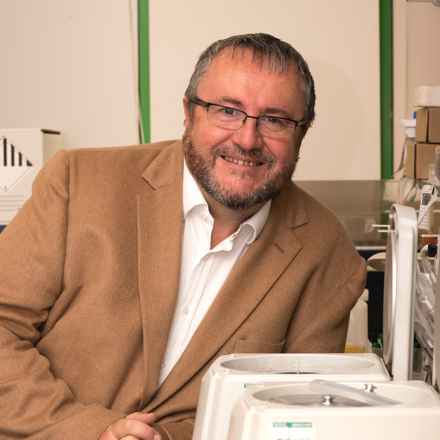 Pete Coffey, a white man in his 60s with grey hair and a beard wearing a brown suit jacket smiling and leaning against lab equipment