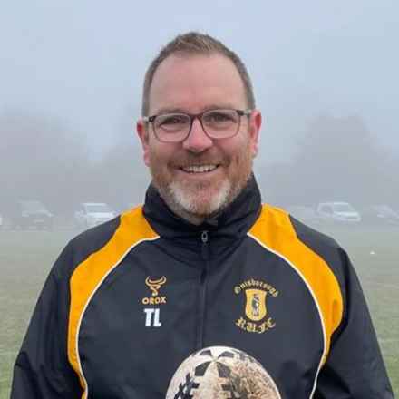 Trevor has short light brown hair and glasses, smiling down the camera outside on a foggy day. He is wearing his rugby club's dark coloured jacket with orange stripes coming down from each shoulder. The jacket includes the Guisborough Rugby Club crest and his initials. The top half of a rugby ball can also be seen which Trevor is holding.