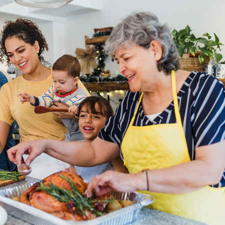 Family Christmas scene. Older woman in apron preparing a cooked turkey, smiling, surrounded by family, including a young girl and a baby being held by his mother who is helping to prepare food