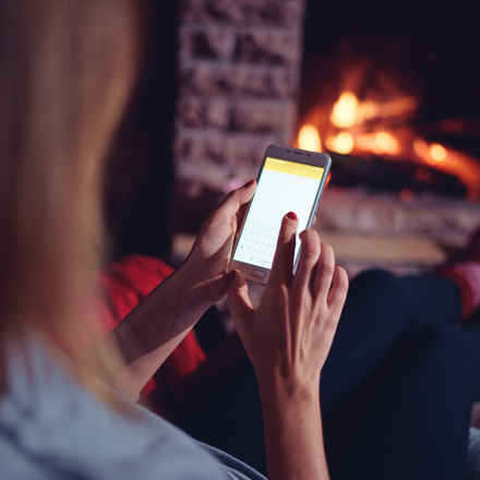 Winter Warmer. Woman on smart phone by cozy fire.
