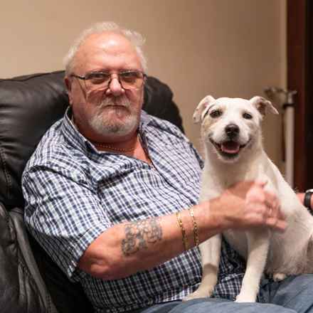 Duncan sitting in his living room. He has short white hair, glasses and is wearing a checked shirt while holding his dog - a smiling white terrier.