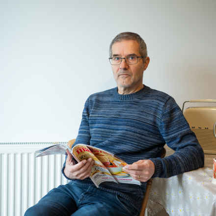 Simon staring at camera, while holding a magazine. A radio and cup of tea sit on the table